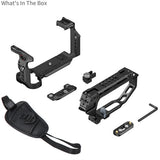 Black camera rig accessory kit laid out on a white background, including a handle grip, mounting brackets, a small Allen wrench, and a strap. Text in the top left reads 'What's In The Box'.