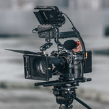 A professional film camera rig with a matte box, monitor, microphone, and tripod, photographed against a blurred outdoor background.