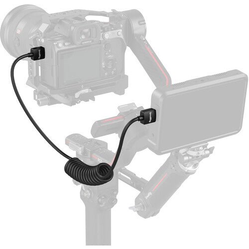 A black coiled cable adapter with two small black connector blocks, shown attached to a camera and gimbal setup on a light gray background.