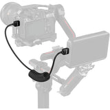 A black coiled cable adapter with two small black connector blocks, shown attached to a camera and gimbal setup on a light gray background.