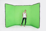 A woman stands in front of a large curved green screen backdrop with a visible black support frame, photographed against a clean white studio background.