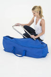 A woman kneels beside a large blue soft-sided storage bag or duffel-style case while holding a metal frame or handle against a clean white studio background.